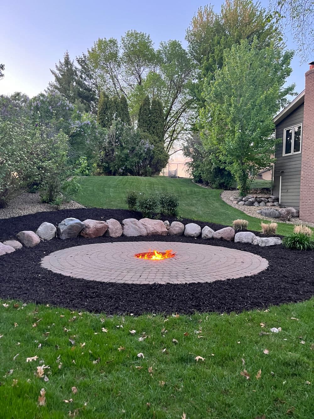 Circular stone fire pit surrounded by landscaped garden and trees in residential backyard.